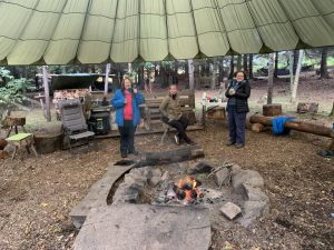 Three female veterans around a fire under a yurt tent holding hot drinks